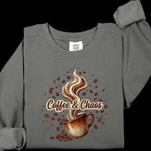 May include: A charcoal gray long-sleeve sweatshirt featuring a graphic of a brown coffee mug with swirling steam. The text "Coffee & Chaos" is printed in brown and white. Scattered brown coffee beans and small heart shapes surround the mug.