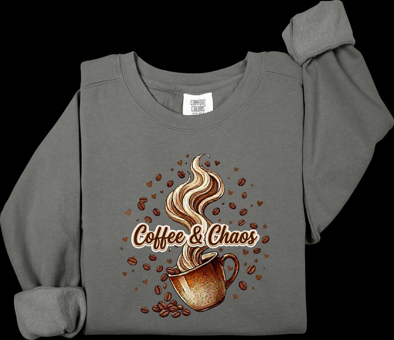May include: A charcoal gray long-sleeve sweatshirt featuring a graphic of a brown coffee mug with swirling steam. The text "Coffee & Chaos" is printed in brown and white. Scattered brown coffee beans and small heart shapes surround the mug.