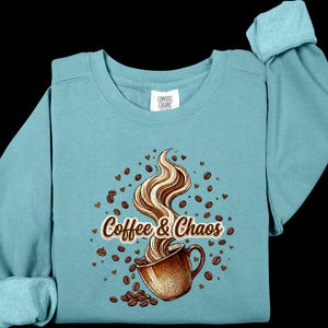 May include: A teal crewneck sweatshirt featuring a graphic of a brown coffee mug with swirling steam, surrounded by scattered coffee beans and small hearts. The text "Coffee & Chaos" is printed in a brown script font across the center of the design.
