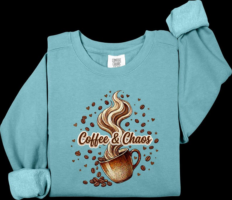 May include: A teal crewneck sweatshirt featuring a graphic of a brown coffee mug with swirling steam, surrounded by scattered coffee beans and small hearts. The text "Coffee & Chaos" is printed in a brown script font across the center of the design.