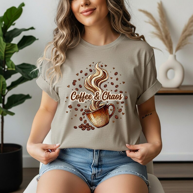 May include: A light beige t-shirt featuring a graphic of a brown coffee mug with swirling steam rising from it. The text "Coffee & Chaos" is printed in a brown script font across the center, surrounded by scattered coffee beans and small hearts.