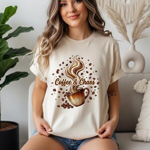 May include: A cream-colored short-sleeve t-shirt featuring a graphic print of a brown coffee mug with swirling steam rising from it. The text Coffee & Chaos is printed in brown script, surrounded by scattered coffee beans and small heart shapes.