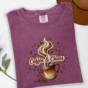 May include: A plum-colored t-shirt featuring a graphic of a brown coffee mug with swirling steam rising from it. The text Coffee & Chaos is printed in a white font with a brown outline. Scattered brown coffee beans and small hearts surround the mug.
