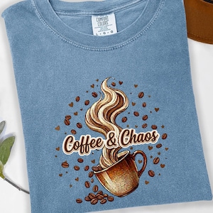 May include: A blue short-sleeve t-shirt with a graphic print of a brown coffee mug with swirling steam, scattered coffee beans, and small hearts. The text "Coffee & Chaos" is printed in brown letters across the center of the graphic design.