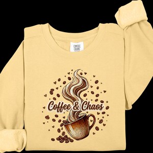 May include: A pale yellow long-sleeve sweatshirt featuring a graphic of a brown coffee mug with swirling steam, surrounded by scattered coffee beans, small hearts, and stars. The text "Coffee & Chaos" is printed in brown with a white outline.