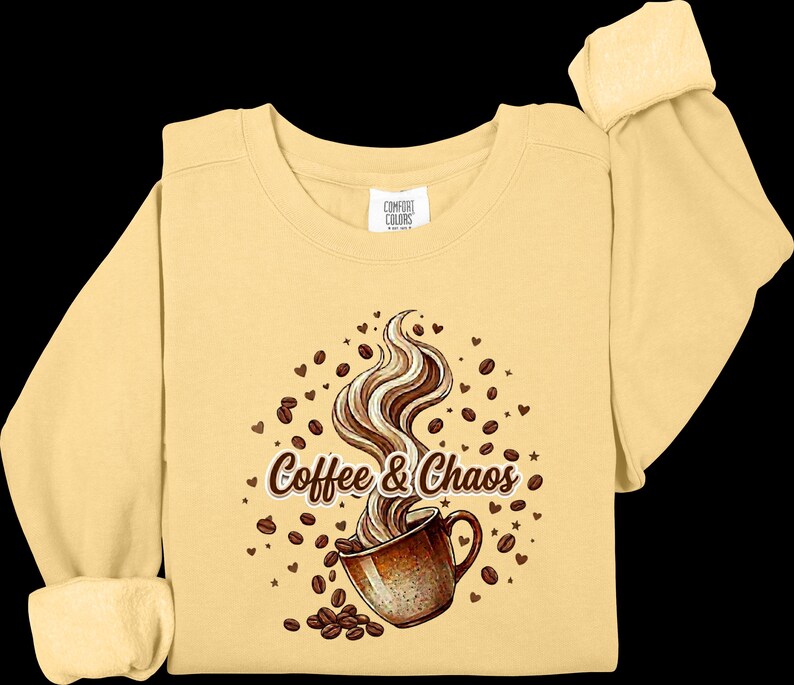 May include: A pale yellow long-sleeve sweatshirt featuring a graphic of a brown coffee mug with swirling steam, surrounded by scattered coffee beans, small hearts, and stars. The text "Coffee & Chaos" is printed in brown with a white outline.