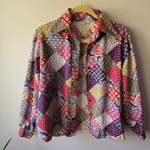 Puede incluir: Una camisa patchwork vintage con un diseño geométrico colorido. La camisa de manga larga presenta una parte delantera abotonada y un cuello ancho. El patrón incluye diseños de cachemira, florales y geométricos en tonos rojos, azules, verdes y amarillos.