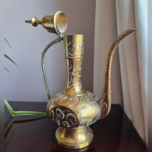 Puede incluir: Una jarra de latón vintage con un pico alargado y una tapa con bisagras. La jarra presenta intrincados patrones florales grabados y un asa curvada. El latón tiene un cálido tono dorado.