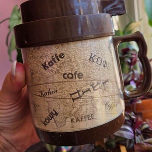 May include: A brown plastic travel mug featuring a beige world map background with the word coffee printed in various languages including Kaffe, cafe, Kahvi, Kaffee, and Japanese characters. The mug has a dark brown lid and a matching handle.