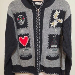 May include: A gray and black zip-up hooded knit sweater featuring embroidered patches of a basket, a red heart, a Christmas tree, and white foliage. The garment has two front pockets, a silver zipper, and a NorthCrest brand label.