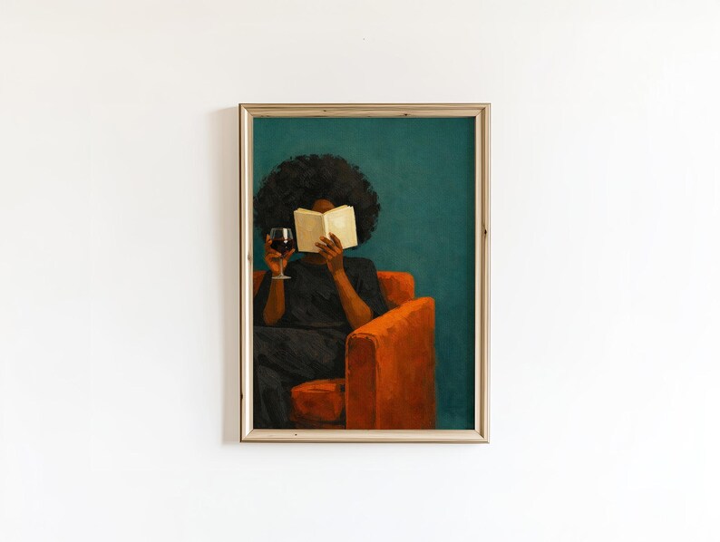 May include: A framed wall art print depicting a person with dark skin and voluminous hair sitting in a bright orange armchair. The person wears a black shirt, holds an open book in front of their face, and holds a glass of red wine in one hand.