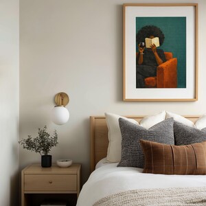 May include: A framed wall art print featuring a painting of a person with dark curly hair sitting in an orange armchair, reading a book while holding a glass of red wine. The artwork is set against a deep teal background and encased in a light wood frame.