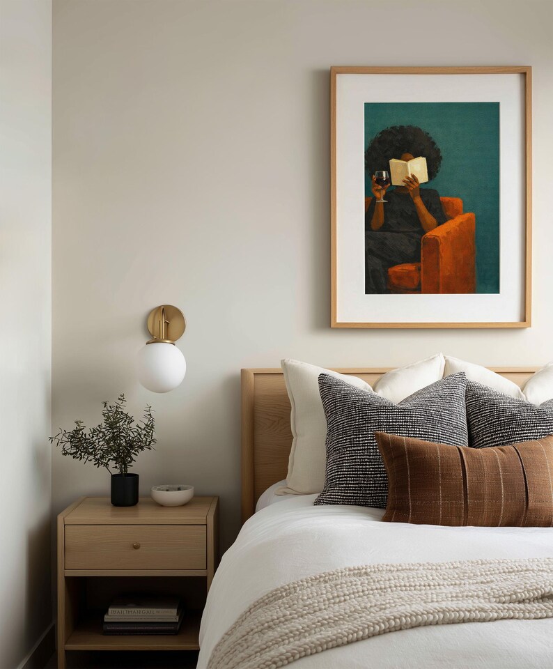 May include: A framed wall art print featuring a painting of a person with dark curly hair sitting in an orange armchair, reading a book while holding a glass of red wine. The artwork is set against a deep teal background and encased in a light wood frame.
