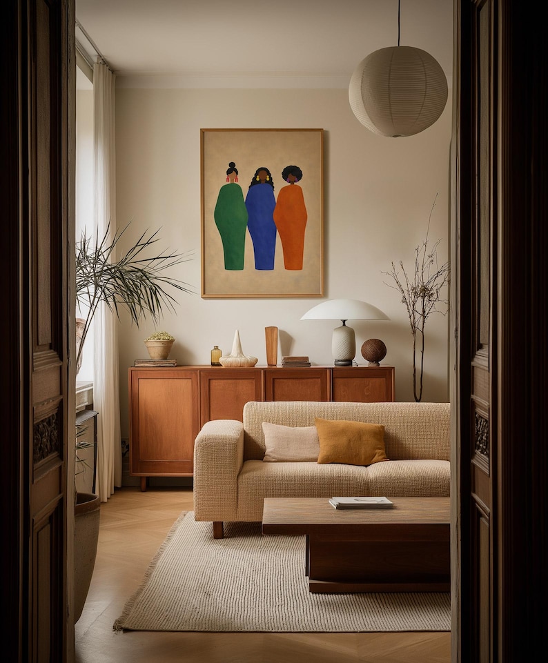 May include: A framed art print featuring three figures in green, blue, and orange robes. The artwork hangs above a wooden cabinet and a beige sofa with throw pillows. A paper lantern and a table lamp are also visible.