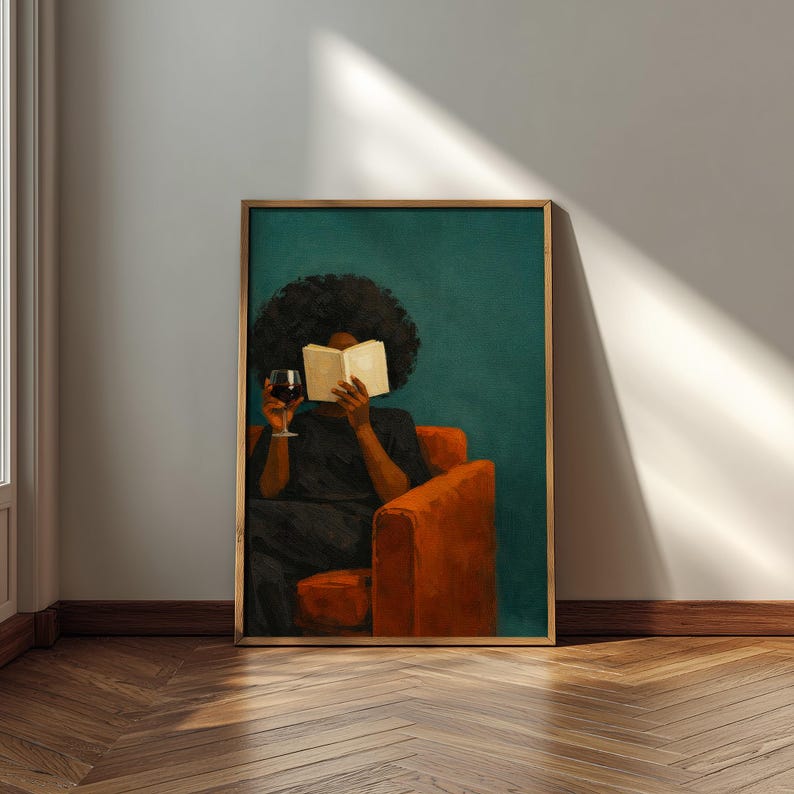 May include: A framed wall art print depicting a person with dark curly hair sitting in an orange armchair, reading an open book while holding a glass of red wine. The background is a solid dark teal color, and the frame is a metallic gold finish.