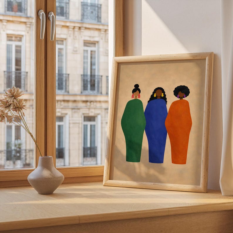May include: A framed art print featuring three stylized figures in green, blue, and orange dresses. The figures have dark hair and are adorned with earrings. The artwork is displayed on a wooden surface near a window.