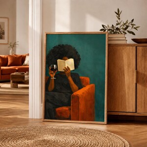 May include: A framed wall art print depicting a person with dark curly hair sitting in an orange armchair. The person is wearing a dark shirt, holding an open book in front of their face, and holding a glass of red wine in their other hand.