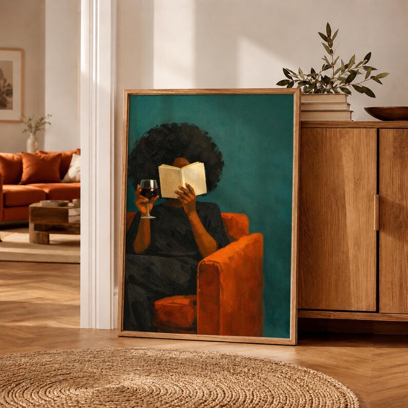 May include: A framed wall art print depicting a person with dark curly hair sitting in an orange armchair. The person is wearing a dark shirt, holding an open book in front of their face, and holding a glass of red wine in their other hand.