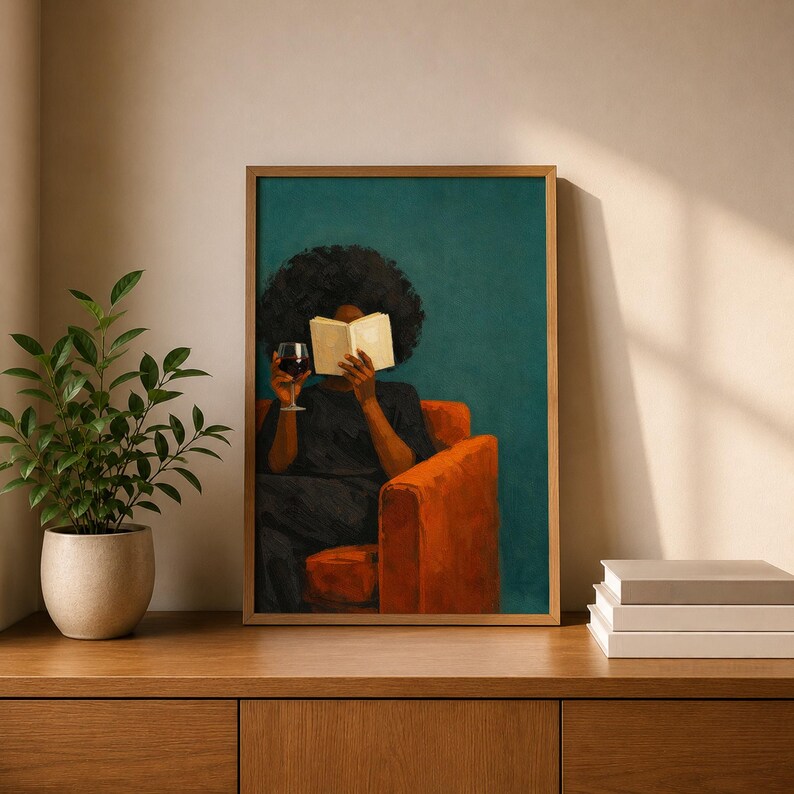 May include: A framed wall art print depicting a person with dark curly hair sitting in an orange armchair, holding an open book in front of their face and a glass of red wine in their other hand, set against a solid dark teal background.