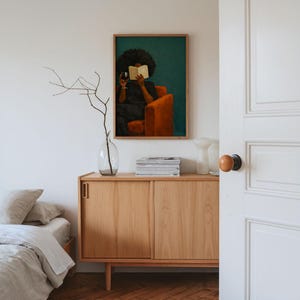 May include: A framed wall art print featuring a painting of a person with dark curly hair sitting in an orange armchair, reading an open book while holding a glass of red wine against a deep teal background, displayed above a light wood cabinet.
