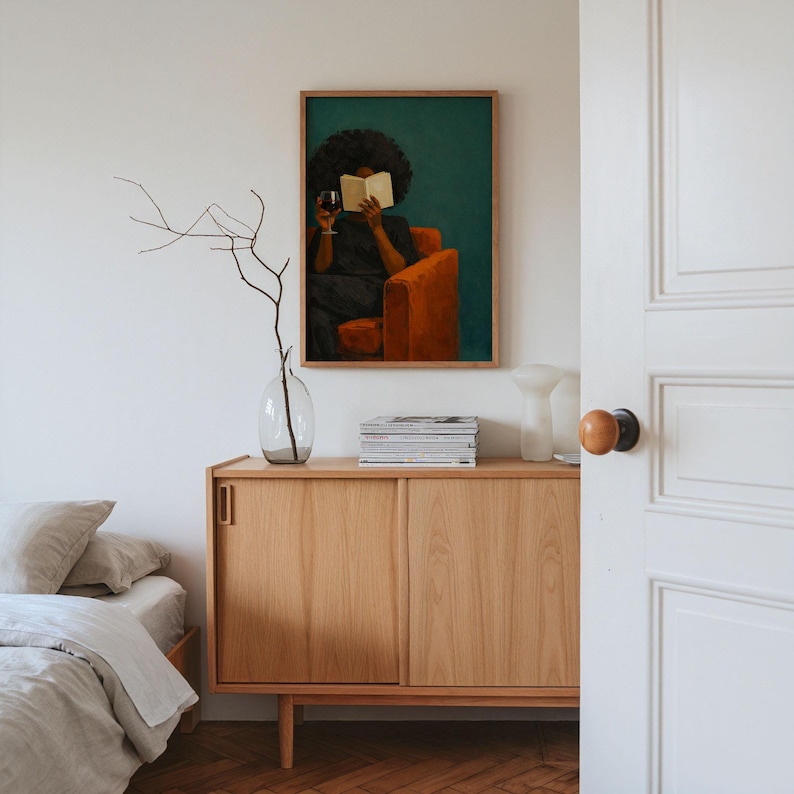May include: A framed wall art print featuring a painting of a person with dark curly hair sitting in an orange armchair, reading an open book while holding a glass of red wine against a deep teal background, displayed above a light wood cabinet.