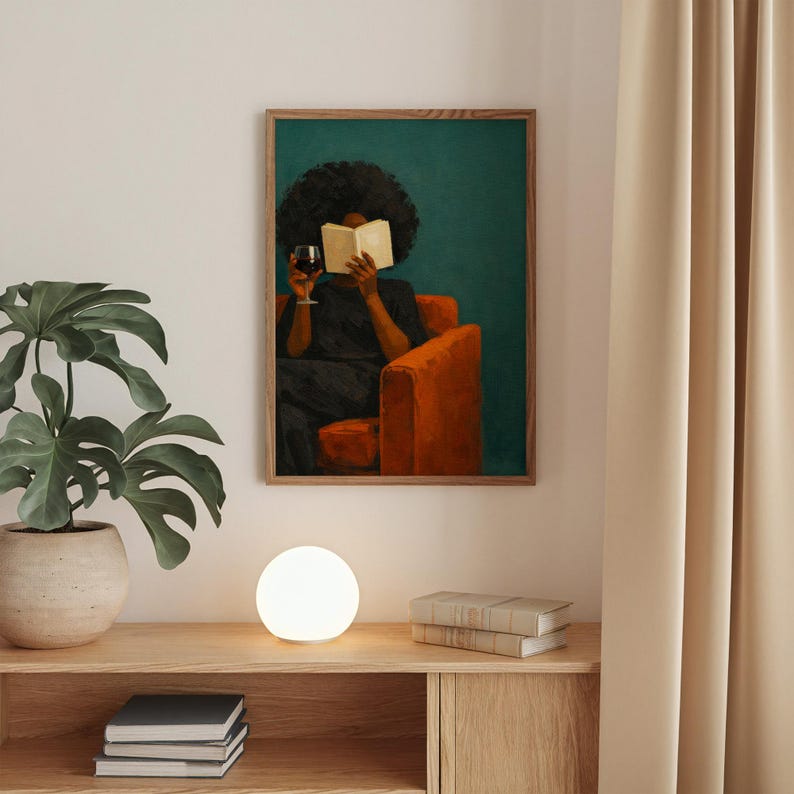 May include: A framed wall art print featuring a person with dark curly hair sitting in an orange armchair, reading an open book while holding a glass of red wine. The background is a solid teal color, and the artwork is displayed in a wooden frame.