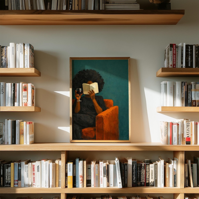 May include: A framed wall art print featuring a person with dark curly hair sitting in a bright orange armchair, reading an open book while holding a glass of red wine. The background is a solid teal color, and the print is displayed on a wall.