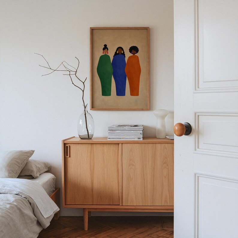 May include: A framed art print featuring three figures in solid green, blue, and orange dresses. The artwork hangs above a wooden cabinet with a glass vase and a stack of magazines. The room has a neutral color palette with a bed visible on the left.