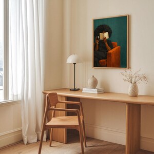 May include: A framed wall art print featuring a person with dark curly hair sitting in an orange armchair, reading a book and holding a glass of red wine against a dark teal background, displayed in a room with a wooden desk and chair.