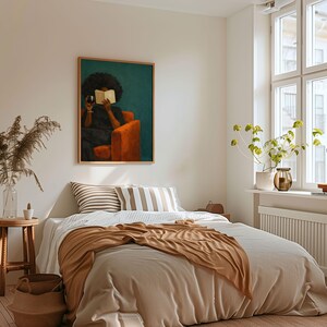 May include: A rectangular wall art print featuring a person with dark curly hair sitting in an orange armchair, holding a book and a glass of red wine against a dark teal background, displayed in a light wood frame above a bed with neutral bedding.