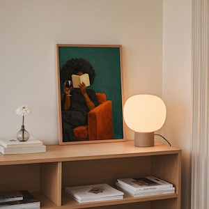May include: A framed rectangular art print featuring a person with dark curly hair sitting in an orange armchair, reading an open book while holding a glass of red wine against a dark teal background, positioned next to a round white table lamp.