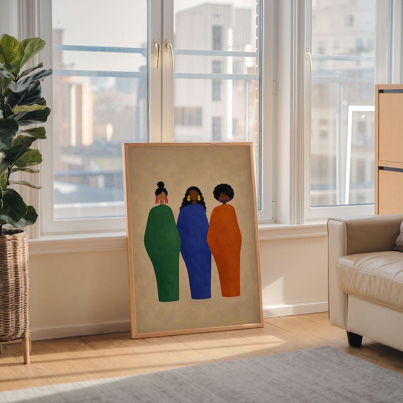 May include: A framed art print featuring three stylized figures in green, blue, and orange dresses. The figures have dark hair and are set against a beige background. The artwork is displayed in a room with a window and a plant.