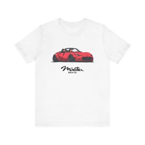 May include: White t-shirt with a red illustration of a Miata MX-5 convertible car. The car is shown from a low angle, with black text reading "Miata MX-5" underneath. The shirt is made of a soft, comfortable material.