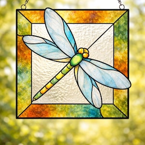 May include: Stained glass dragonfly suncatcher with a square frame. The dragonfly has light blue wings and a yellow and green body. The frame has a mix of orange, green, and yellow colors. It hangs from a metal chain.