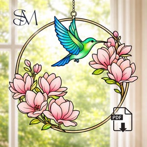 Puede incluir: Una corona de flores y colibríes de vidriera. El colibrí tiene alas azules y verdes y un pico largo, flotando cerca de flores rosas. La corona es de color dorado y cuelga de una cadena. El fondo es una vista borrosa de una ventana.