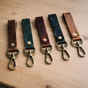 May include: Five leather keychains in various colors: brown, green, burgundy, black, and tan. Each keychain features a brass-colored clasp and a matching rivet. The keychains are arranged on a light-colored wooden surface.