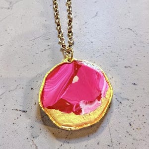 May include: A gold-toned necklace featuring a pendant with a vibrant, abstract design. The pendant has a gold-colored border and a swirling pattern of pink, red, and white hues. The chain is also gold-colored.