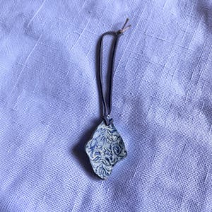 May include: A handmade ceramic pendant with a blue and white floral design. The pendant is diamond-shaped and hangs from a dark gray cord. The background is a light purple linen fabric.