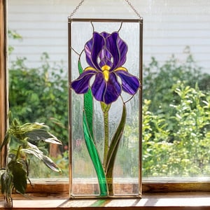 Puede incluir: Panel de vidriera con una flor de iris morada con detalles amarillos. El diseño está en un fondo transparente, enmarcado por un borde metálico. Hojas y tallo verdes completan el diseño floral. El panel cuelga de una cadena.