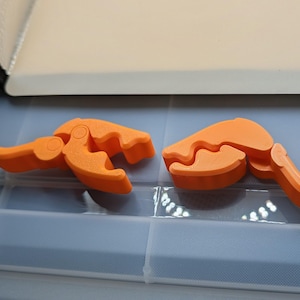 May include: Two orange plastic clamps are displayed. Each clamp has a hinged design with a curved gripping surface and a handle. The clamps are resting on a light blue surface.