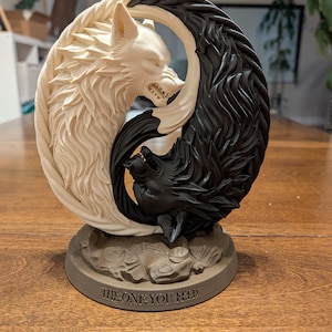 Könnte beinhalten: Skulptur mit zwei Wölfen in einem Yin-Yang-Design. Ein Wolf ist cremefarben, der andere schwarz. Der Sockel ist braun und trägt den Text "THE ONE YOU FEED". Die Wölfe knurren, mit detaillierten Fellstrukturen.