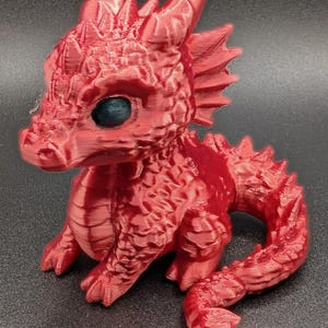 Könnte beinhalten: Eine rote, 3D-gedruckte Drachenfigur mit detaillierten Schuppen, Hörnern und einem eingerollten Schwanz. Der Drache hat eine strukturierte Oberfläche und dunkelblaue Augen, was ihm ein Fantasy-Thema verleiht. Die Figur sitzt.