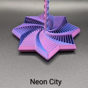May include: A 3D-printed, multi-layered, star-shaped object in shades of purple and blue, with a central, spiraled column. The base has a geometric design, and the text "Neon City" is printed at the bottom. The object is set against a dark background.