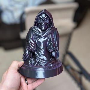 May include: A dark, glossy sculpture of a hooded skull figure. The figure is in a seated pose, with the skull's face and hands visible. The sculpture has a circular base and is held by a hand.