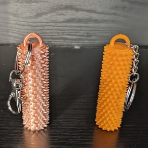 May include: Two textured keychains. One is copper-colored with a silver clasp and ring. The other is orange with a silver chain and ring. Both have a cylindrical shape with a spiky surface.