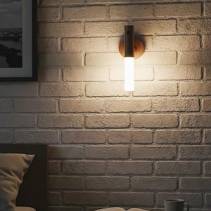 Peut inclure: Applique murale moderne avec une base en bois et un luminaire cylindrique et lumineux, fixée sur un mur de briques blanches. La lumière chaude éclaire le mur, une table de chevet et un cadre photo.