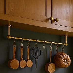Könnte beinhalten: Eine Küchenszene mit einer Messing-Hängestange und verschiedenen Küchenutensilien. Holzlöffel, eine Schere und ein dekorativer Kürbis hängen an der Stange. Die Stange ist unter einem Holzschrank montiert.