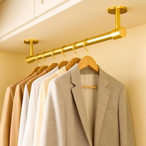 Puede incluir: Una barra de ropa dorada, montada en el techo, con varias perchas de madera que sostienen varias chaquetas y camisas. Las chaquetas y camisas son de colores neutros, incluyendo beige, blanco y gris. La barra está fijada al techo con soportes circulares.