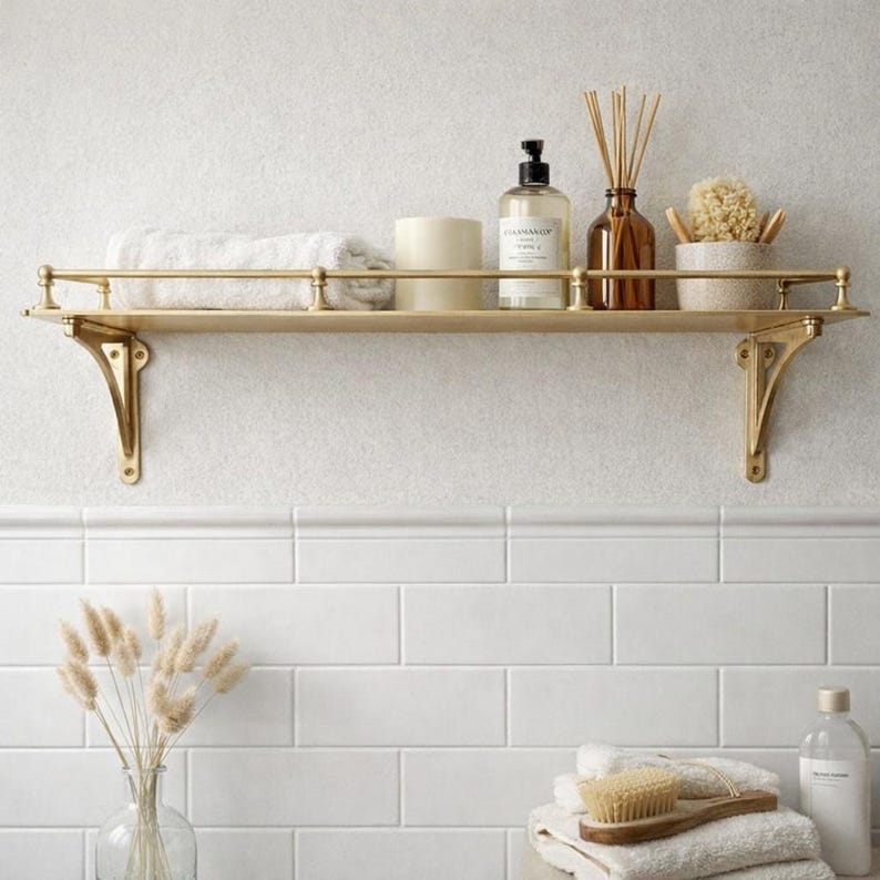 May include: A gold-toned metal wall shelf with a protective railing and two decorative brackets. The shelf holds a folded white towel, a cylindrical cream-colored container, a clear pump bottle with a label, a brown glass bottle with reed diffusers.