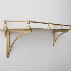 May include: A wall-mounted shelf crafted from brass with a metallic gold finish. The shelf features a flat rectangular surface supported by two decorative triangular brackets and includes a protective railing along the back and sides with four posts.